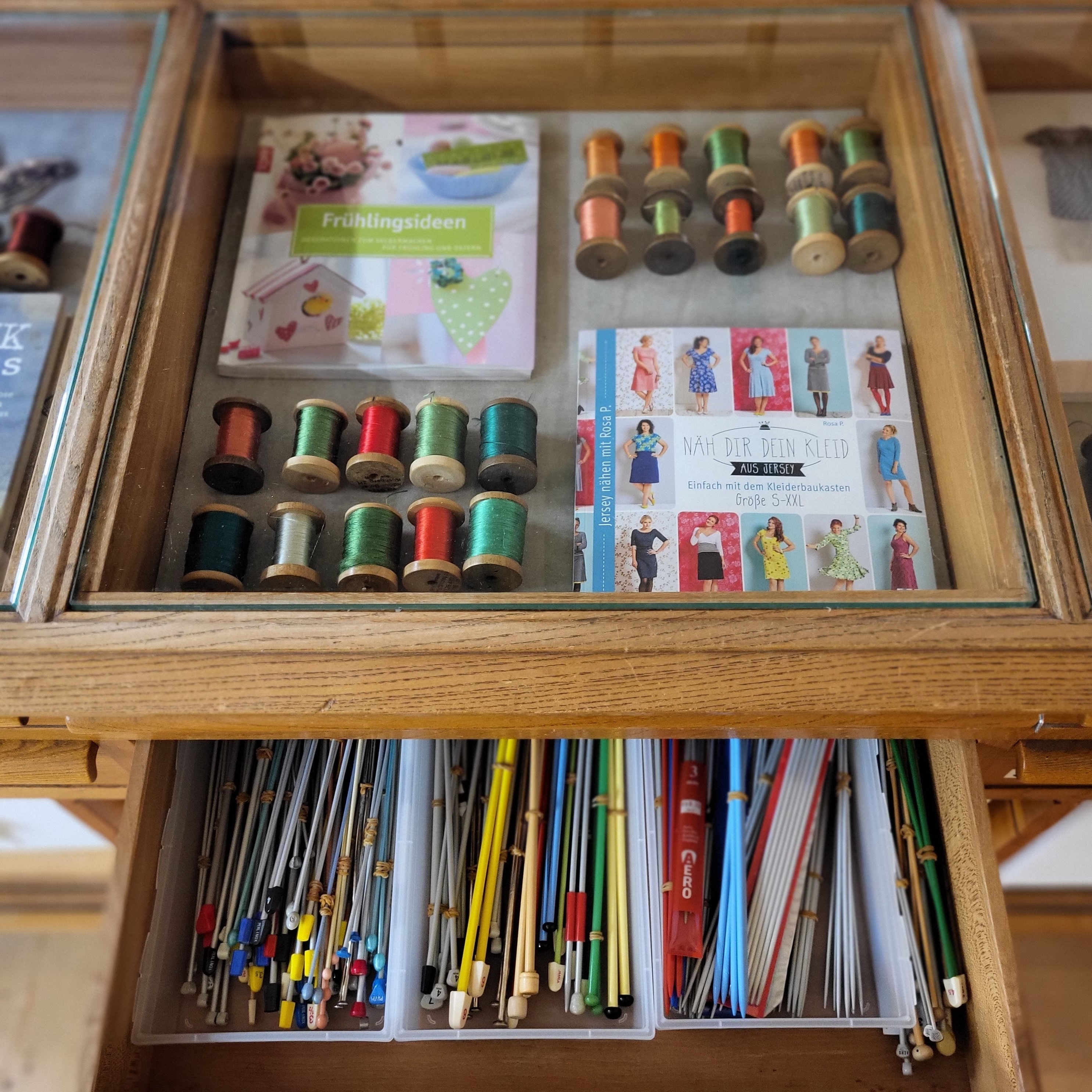 Eine hölzerne Glasvitrine mit Handarbeitsbüchern und bunten Garnrollen über einer geöffneten Schublade voller Stricknadeln