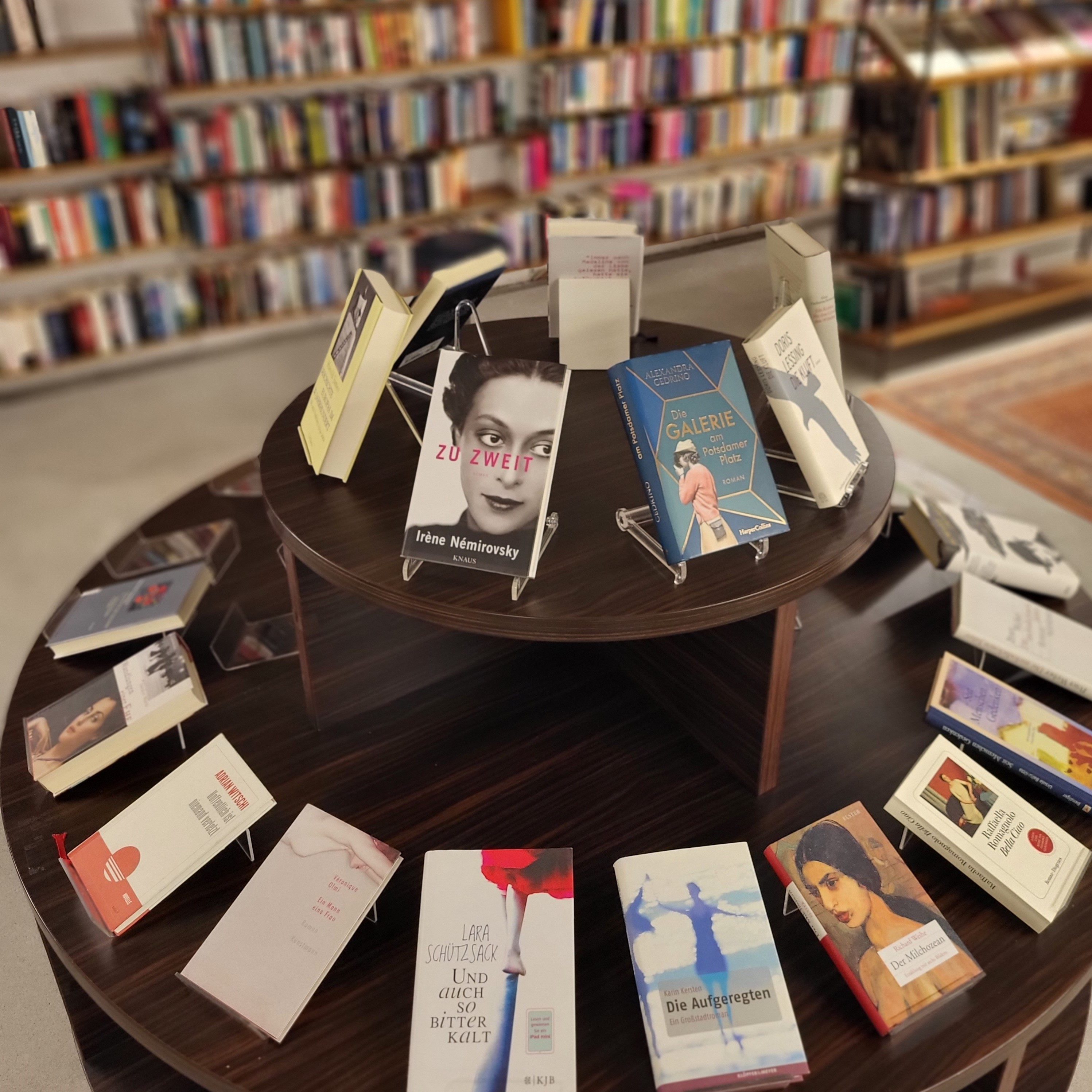 Ein runder, zweistufiger Holztisch in einer Buchhandlung, auf dem verschiedene Romane auf Buchaufstellern präsentiert werden.