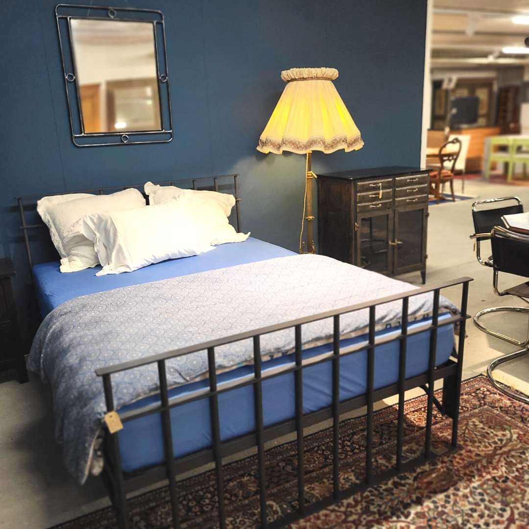 Schlafzimmer-Ausstellung mit einem Metallbett mit blauer Bettwäsche, einer eingeschalteten Stehlampe und einem kleinen Schrank vor einer dunkelblauen Wand.