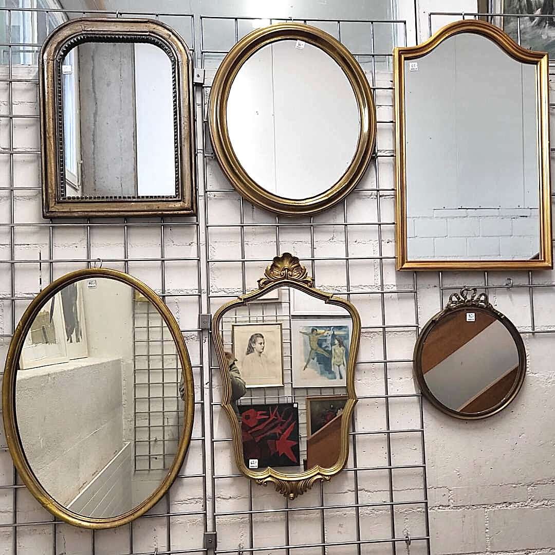 Sechs verschiedene Vintage-Spiegel in unterschiedlichen Formen mit verzierten Goldrahmen, die nebeneinander an einer Metallgitterwand hängen.