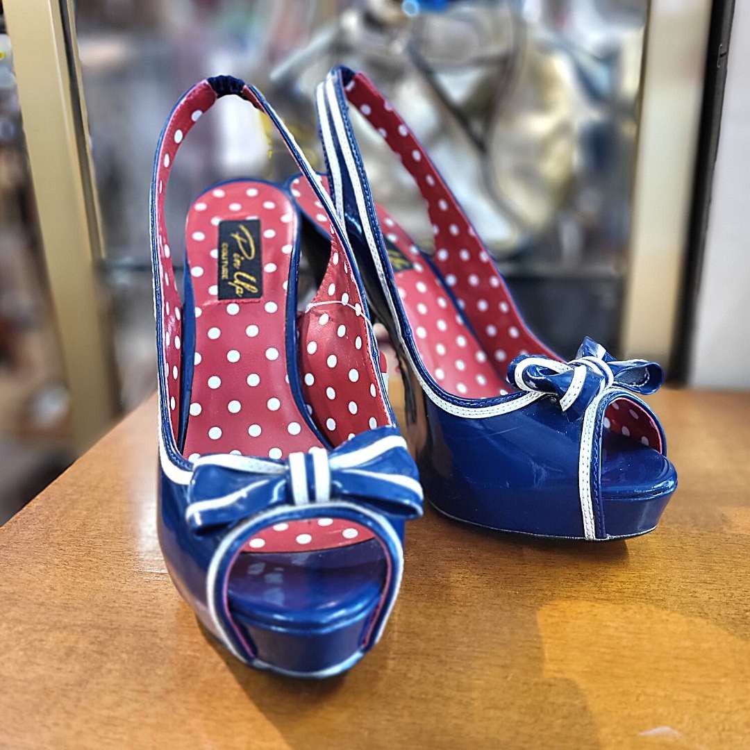 Ein Paar blaue Lack-Slingback-Pumps im Retro-Pin-up-Stil mit hohem Absatz und Plateau, verziert mit einer blau-weissen Schleife an der offenen Zehenpartie (Peep-Toe) und einem rot-weiss gepunkteten Innenfutter samt Markenlabel „Pinup Couture“, präsentiert auf einer braunen Holzoberfläche.