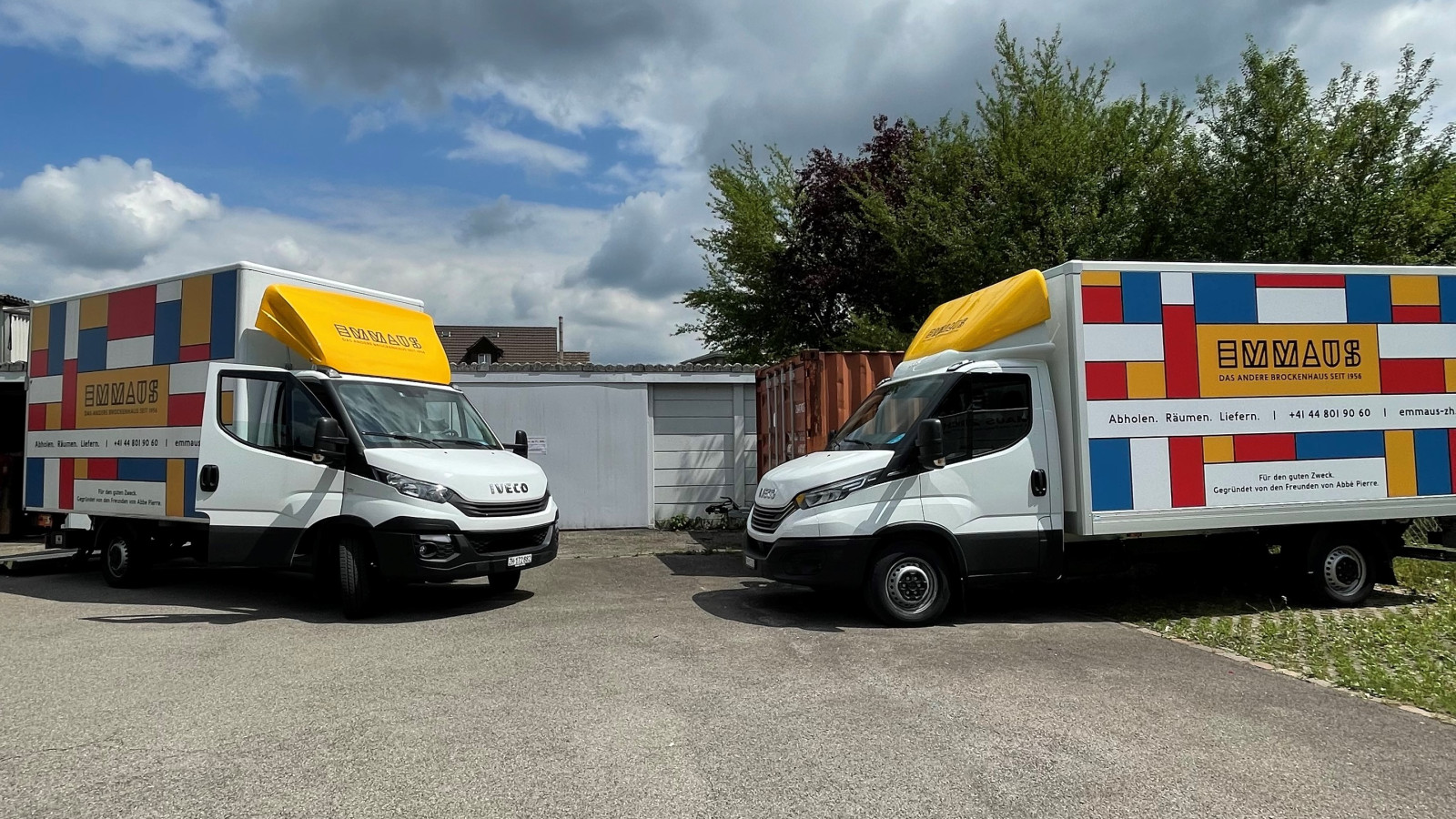 Zwei weisse Lieferwagen der Marke Iveco mit grossen Kofferaufbauten, die nebeneinander auf einem asphaltierten Platz vor Garagentoren und gr&uuml;nen B&auml;umen unter einem leicht bew&ouml;lkten Himmel parken. Die Lkw-Aufbauten und die Dachspoiler sind in einem auff&auml;lligen, bunten Blockmuster im Mondrian-Stil mit roten, blauen und gelben Fl&auml;chen gestaltet und tragen den gelb hinterlegten Schriftzug &bdquo;EMMAUS &ndash; DAS ANDERE BROCKENHAUS SEIT 1994&ldquo;.