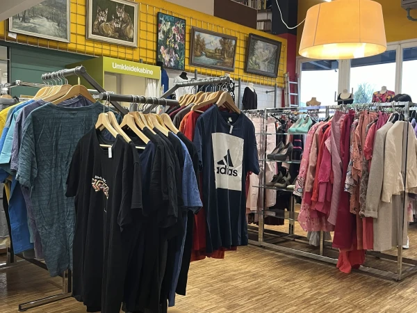 A well-lit secondhand store or boutique interior, featuring several clothing racks filled with various garments on wooden hangers. In the left foreground, a prominent circular rack displays dark t-shirts and short-sleeved tops, including a dark blue shirt with a large Adidas logo. To the right, another rack holds a vibrant selection of women's apparel in shades of pink, red, and beige, alongside shelves displaying high-heeled shoes. The shop has light-colored parquet flooring, a bright yellow upper wall adorned with several framed paintings, a green changing stall labeled "Umkleidekabine," and a large, cylindrical beige lampshade hanging from the ceiling.
