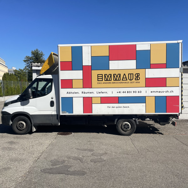 Ein wei&szlig;er Lieferwagen von Emmaus Z&uuml;rich mit farbenfrohem, an Mondrian angelehntem Muster und Beschriftung parkt an einem sonnigen Tag auf einem Asphaltplatz.
